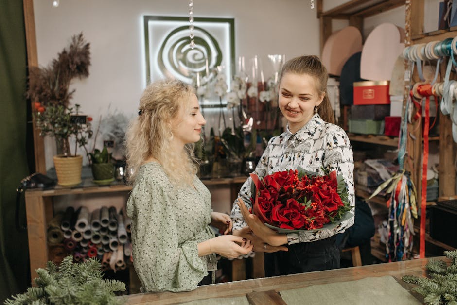 A young woman with light brown hair tied back, dressed in a patterned blouse, is handing a bouquet of fresh red roses and greenery to an older woman with curly blonde hair wearing a light green floral