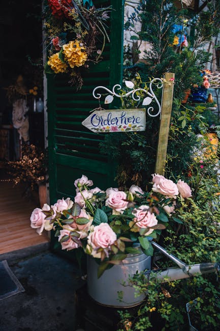 A decorative display at a flower shop featuring a pastel pink rose bouquet in a silver watering can, positioned near a green wooden shutter adorned with hanging floral arrangements. The roses have lus