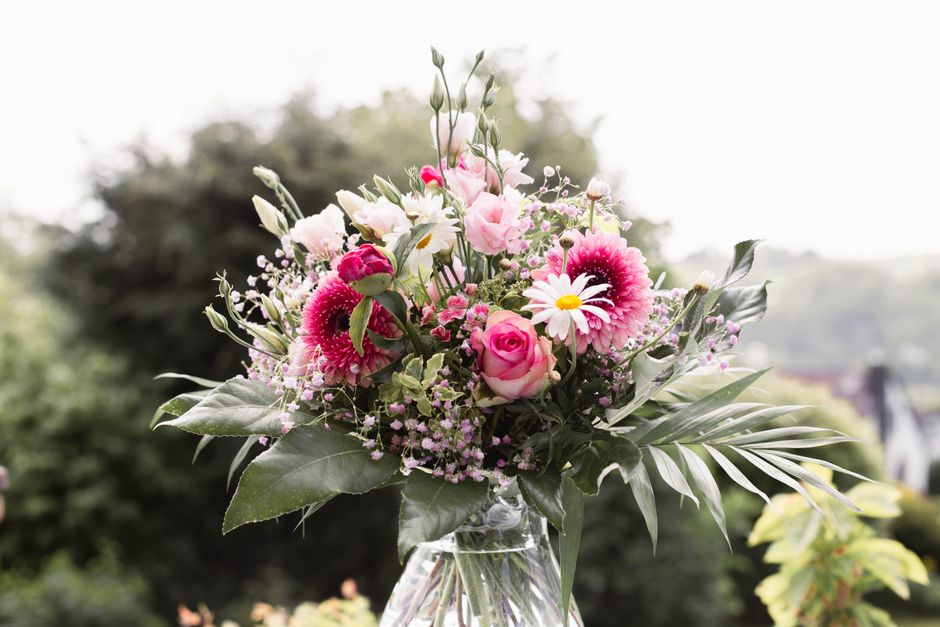 A floral bouquet featuring pink roses, white daisies with yellow centers, and vibrant pink gerbera daisies arranged with lush green foliage and delicate filler flowers. The flowers are set in a clear 