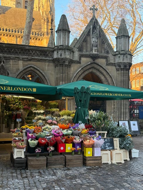 A busy outdoor flower stall set against the historic architecture of a Gothic-style church with pointed arches and ornate stonework. The stall, managed by flowershitchin.co.uk, features an array of co