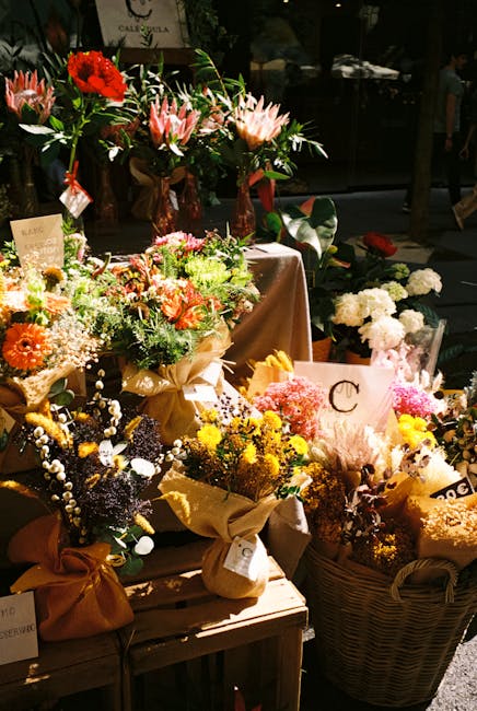 A display of various floral arrangements featuring a mix of fresh flowers, including vibrant orange gerbera daisies, pink proteas, white baby's breath, and yellow chrysanthemums, arranged in wrapped b